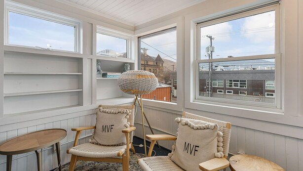 A cozy room with two chairs labeled "MVF," a small table, and a floor lamp. Large windows offer views of buildings outside.