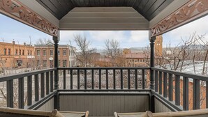 A covered balcony with a wooden railing and two chairs, overlooking a street with buildings and bare trees.
