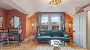 A living room with a teal sofa, a glass coffee table, a round mirror, a desk with a chair, and two floor lamps.