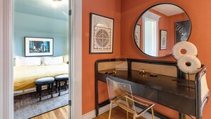 A bedroom with a bed, two stools, a desk, a mirror, and wall art.