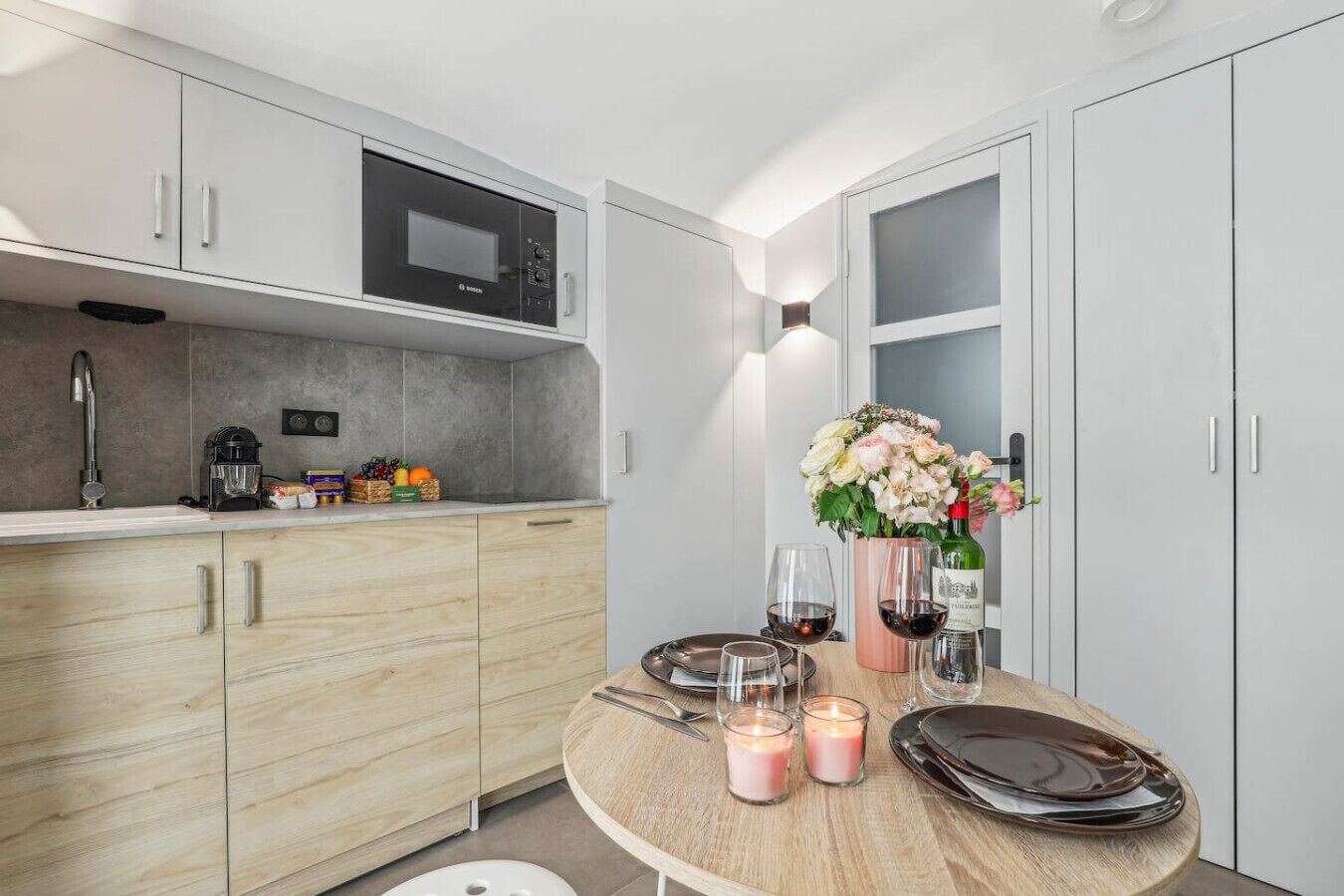 A modern kitchen with a built-in microwave, a coffee machine, and a fruit basket. There is a dining table set with plates, glasses, and candles.