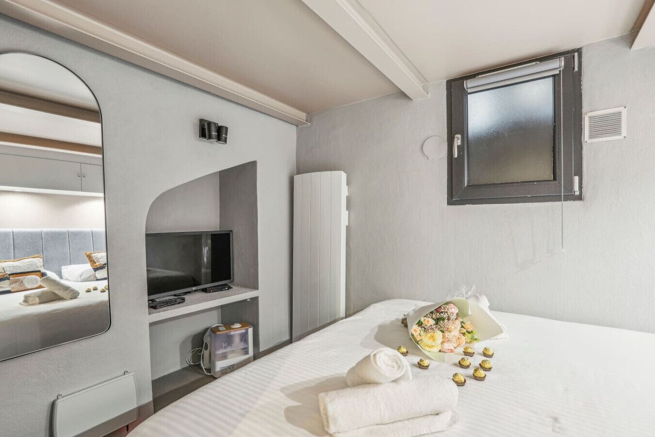 A hotel room with a bed, a TV, a mirror, and a window.