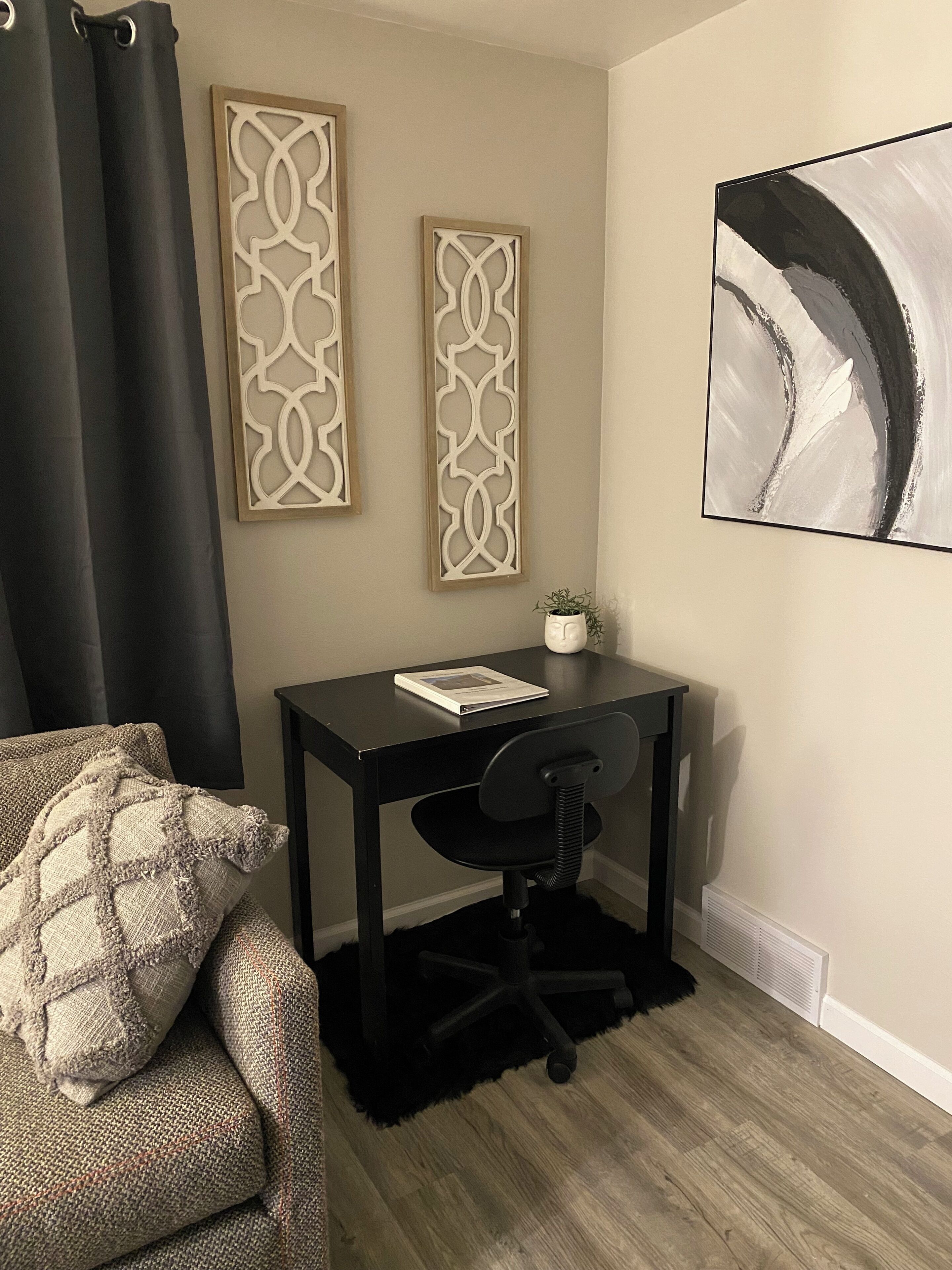 A room with a black desk, a chair, a sofa with a patterned throw, two decorative panels, and a large abstract painting.