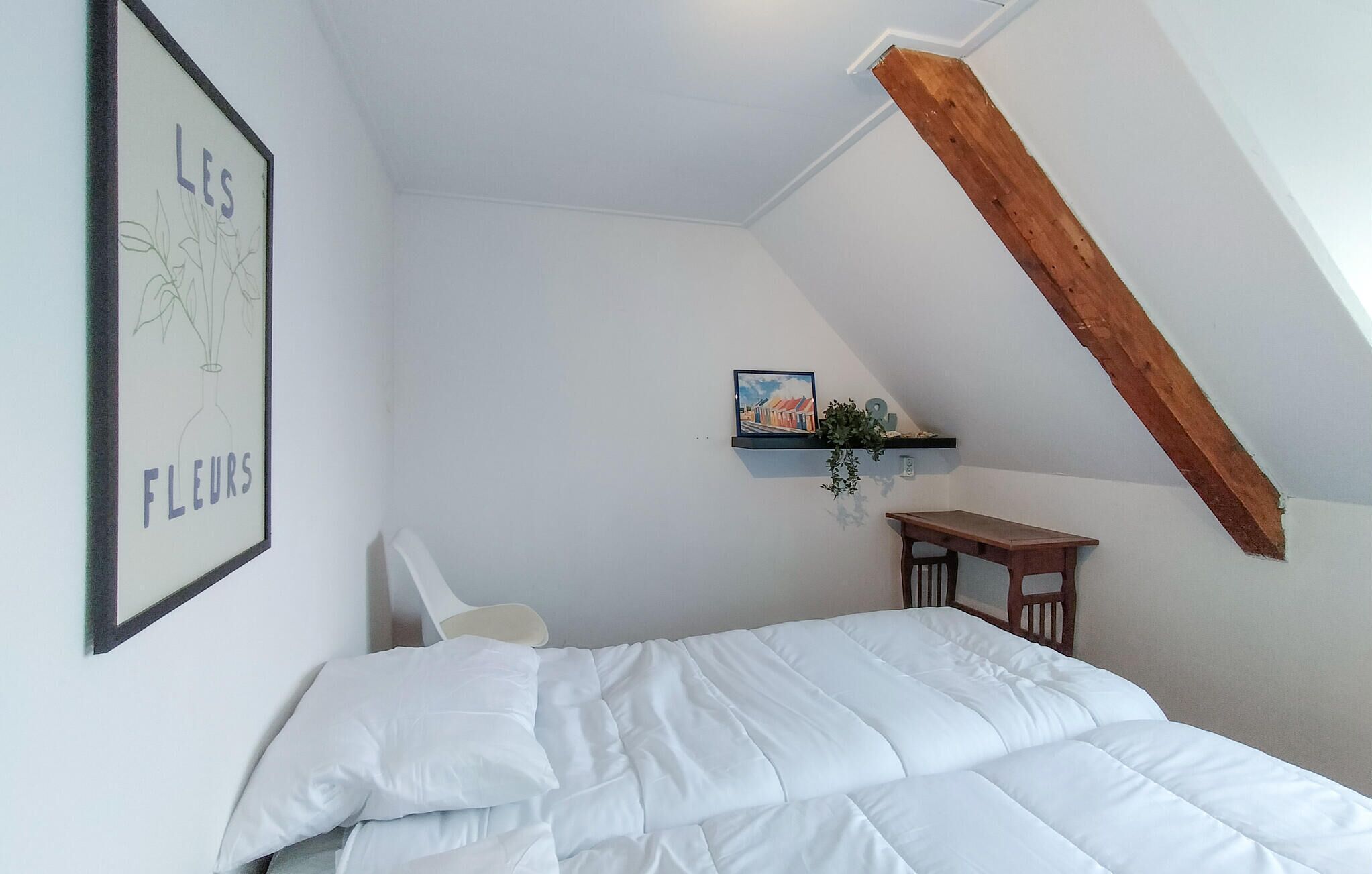 A bedroom with a sloped ceiling, a bed with white bedding, a framed picture of flowers, a small table, and a shelf with a plant.
