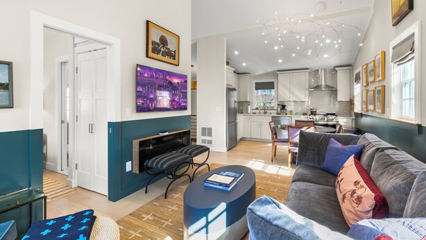 Una sala de estar moderna con un sofá gris, una mesa de centro y una chimenea. Se observa una cocina al fondo con armarios blancos y una campana extractora de acero inoxidable.