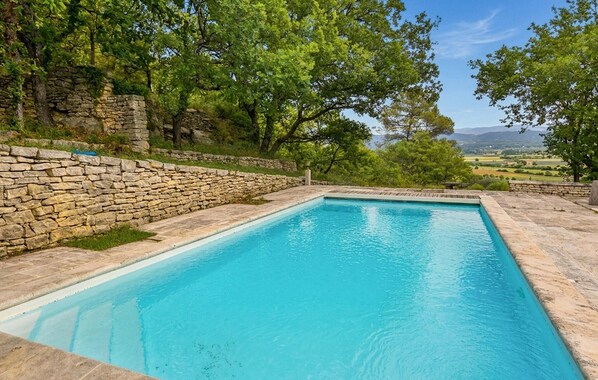 Une piscine rectangulaire à l’eau d’un bleu cristallin, entourée de murs de pierre et d’arbres verdoyants.