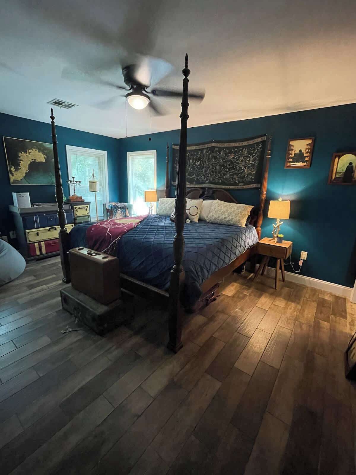 A four-poster bed with a ceiling fan, a wooden floor, and a window with blinds.