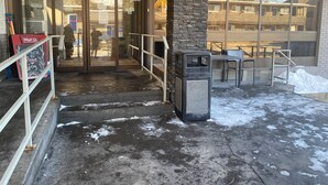 A building entrance with glass doors, a stone pillar, a trash bin, and a seating area with tables and chairs.
