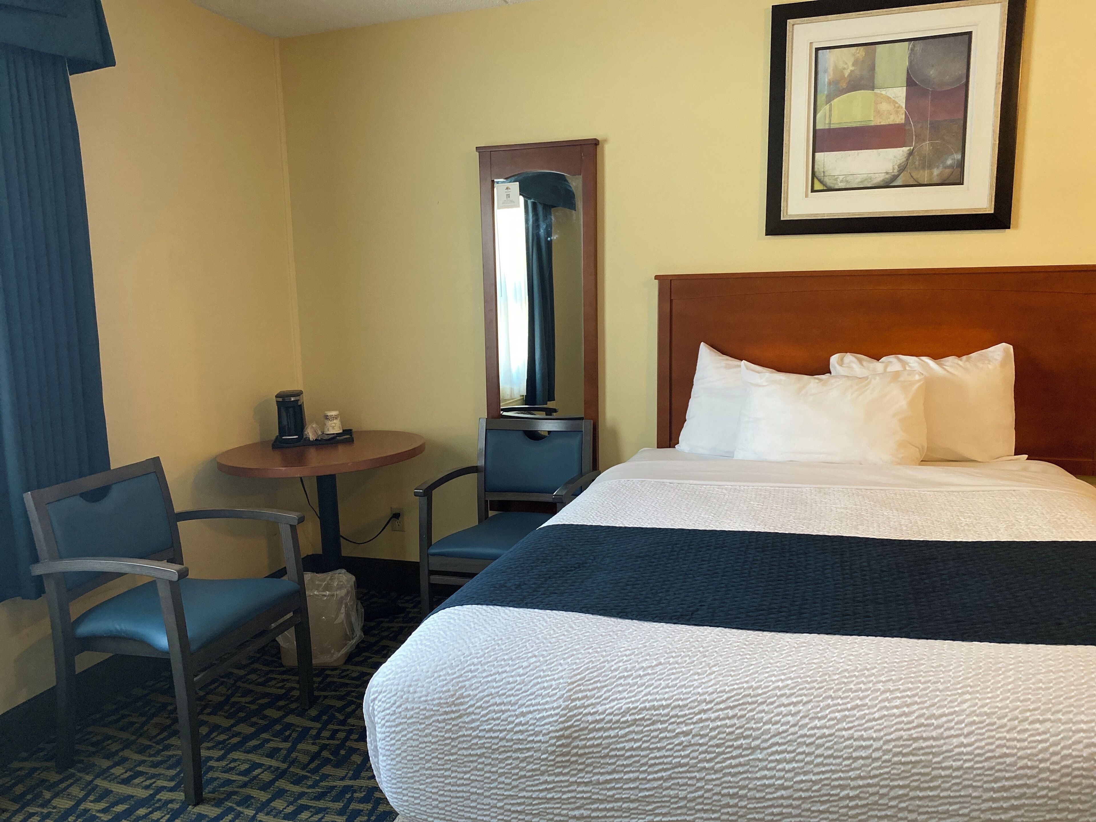 A hotel room with a large bed, a small table with a coffee maker, a chair, and a mirror.