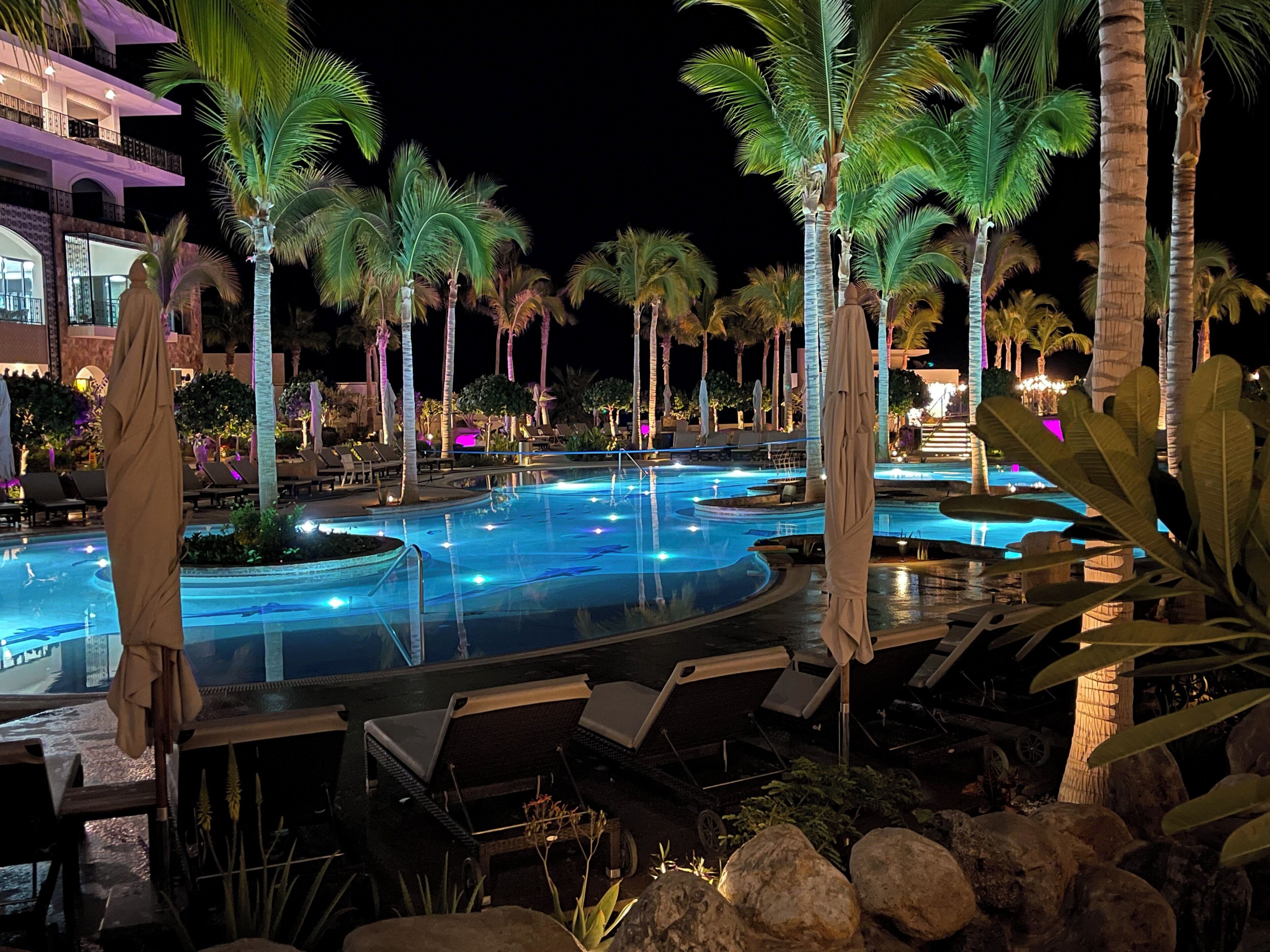 Een zwembadcomplex van een resort bij nacht, met meerdere zwembaden, palmbomen, ligbedden en parasols.