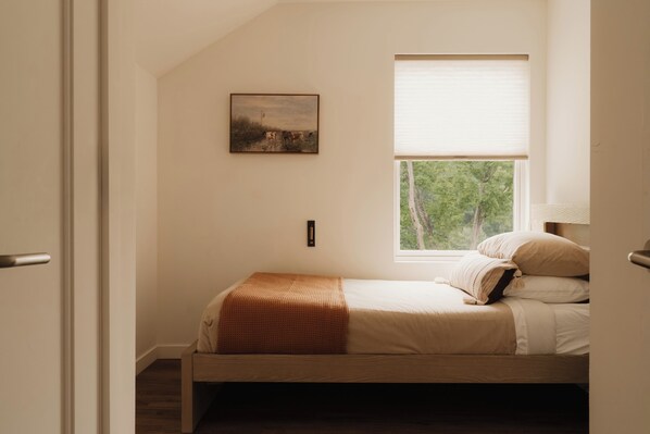 A bedroom with a single bed, a window with a view of trees, and a painting on the wall.