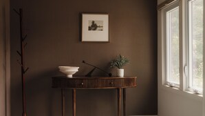 Een hoekzaak met een donkere wand, een ingelijst beeld, een houten console met een schaal en een plant, en een vloerlamp.