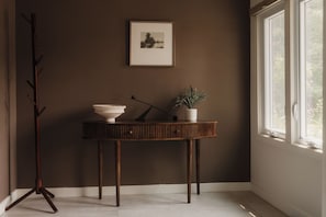 Una habitación en esquina con una pared oscura, un cuadro enmarcado, una consola de madera con un cuenco y una planta, y una lámpara de pie.