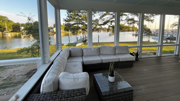 A covered outdoor seating area with a large sectional sofa and a small table, overlooking a lake with boats and greenery. - Stunning Chesapeake Waterfront Home (Oxford)