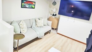 A living room with a sofa, a small side table, a TV mounted on the wall, and a decorative wall hanging.