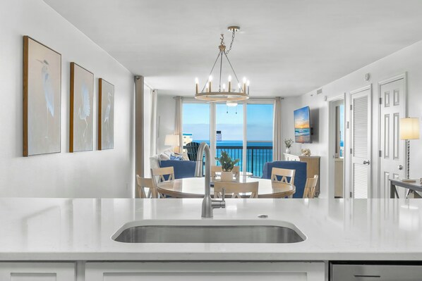 A modern kitchen with a central island, a dining area with blue chairs, and a view of the ocean through large windows.