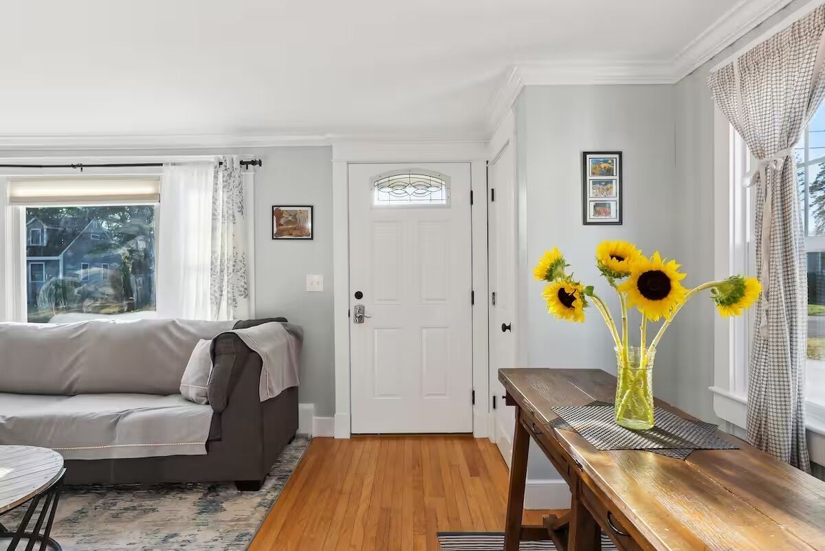 A living room with a sofa, a wooden table with a vase of sunflowers, a white door, and a window with curtains.