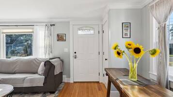 A living room with a sofa, a wooden table with a vase of sunflowers, a white door, and a window with curtains.
