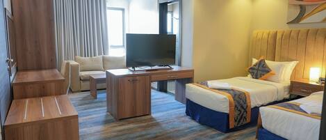 Ein Hotelzimmer mit einem großen Bett, einem kleineren Bett, einem Schreibtisch mit Fernseher, einem Sessel und einem Kleiderschrank.