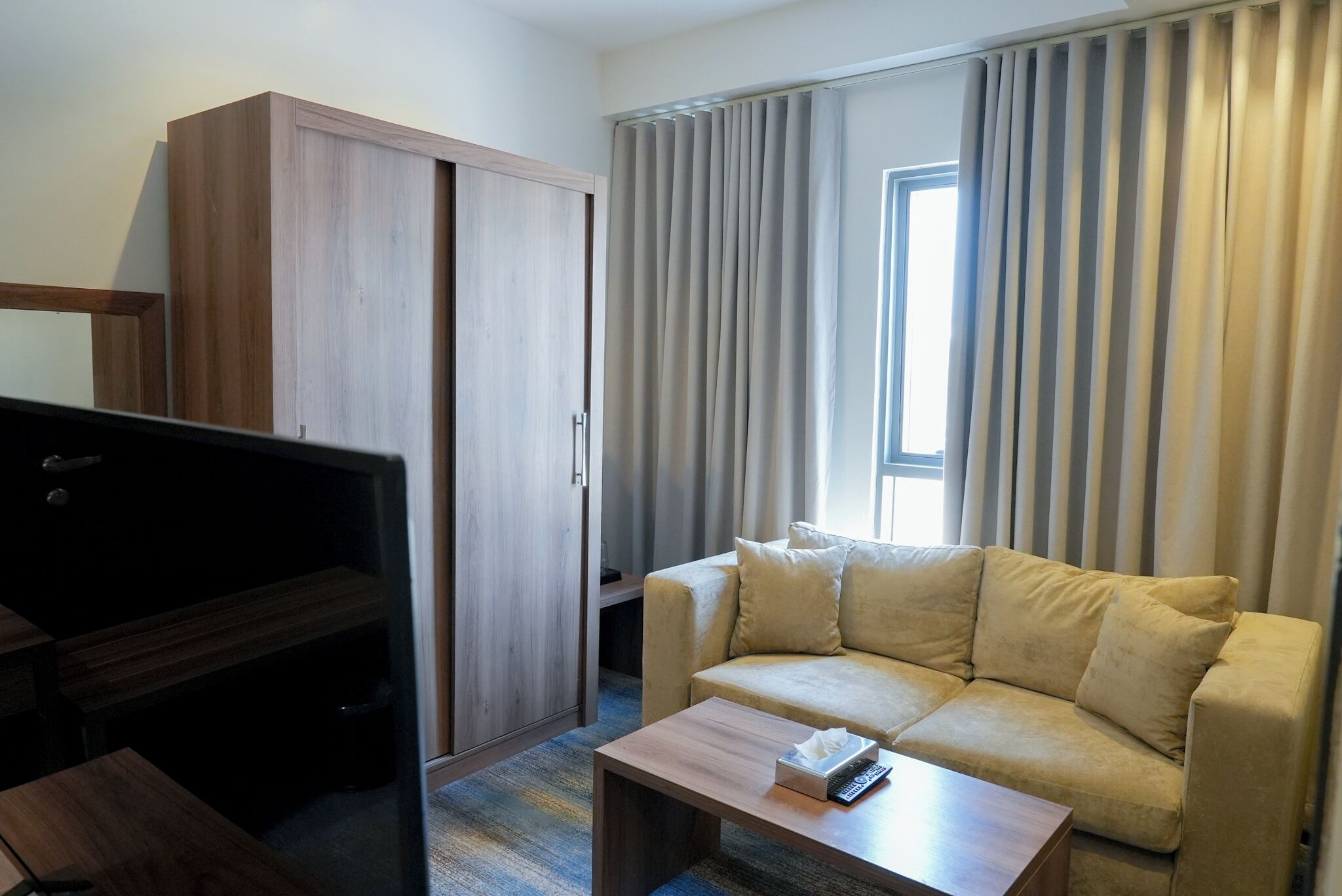 Ein Hotelzimmer mit einer Couch, einem Fernseher, einem Holzschrank und einem Fenster mit Vorhängen.