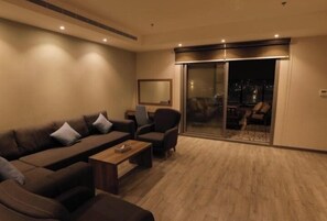 Una sala de estar moderna con sofá, sillones y una mesa de centro. Hay un ventanal corredizo que da a un balcón con vistas a la ciudad.