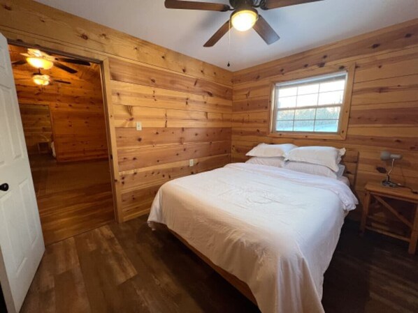 Un dormitorio con paneles de madera, una cama, un ventanal, un ventilador de techo y una mesita de noche pequeña.
