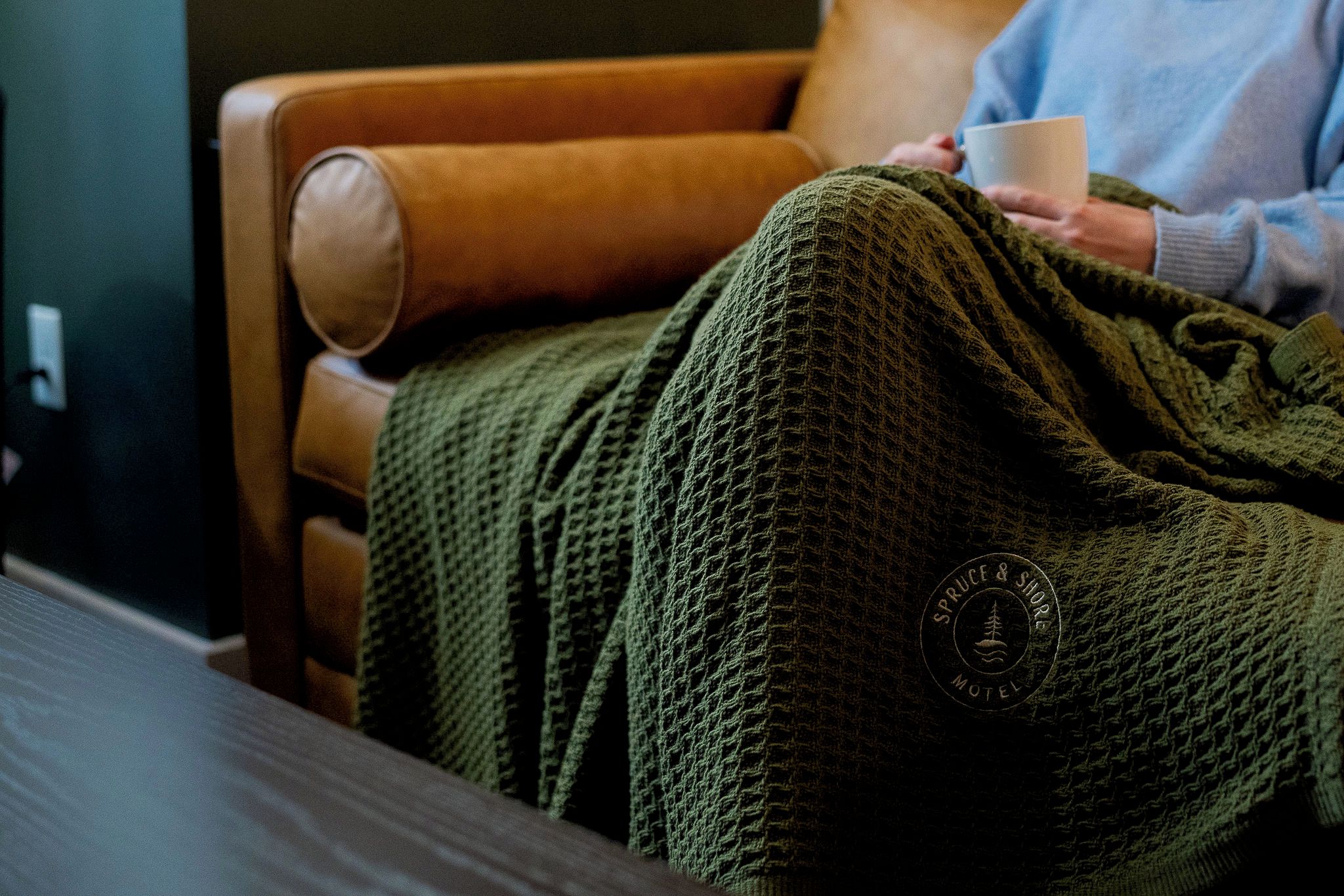 Eine Person sitzt in einem Ledersessel, bedeckt mit einer grünen Strickdecke, und hält eine weiße Tasse.