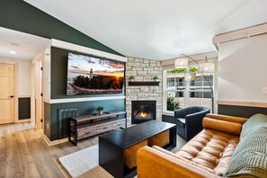 Een moderne woonkamer met een stenen openhaard, een grote flatscreen televisie aan de wand, een comfortabele leren bank en een houten salontafel.
