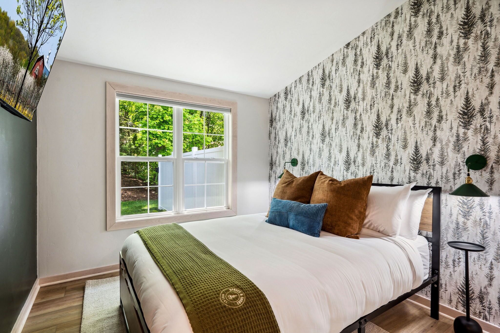 Une chambre avec une grande fenêtre, un lit recouvert de plusieurs oreillers, un plaid vert et un mur orné d’un motif de forêt.