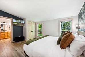 Quarto com uma cama grande, piso de madeira, um quadro e janela com vista para árvores.