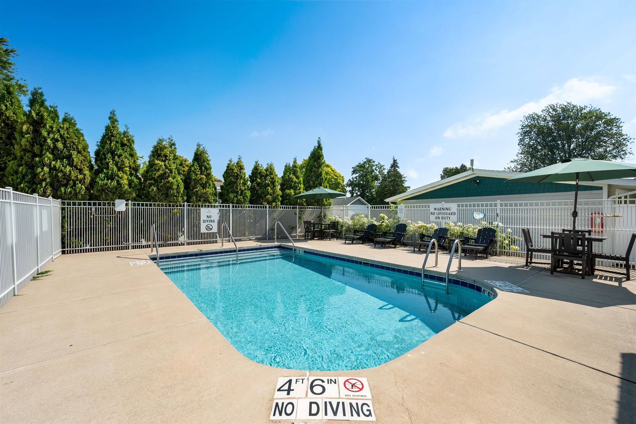 A rectangular pool with a no diving sign at the bottom, surrounded by a white fence, and a patio area with outdoor furniture.