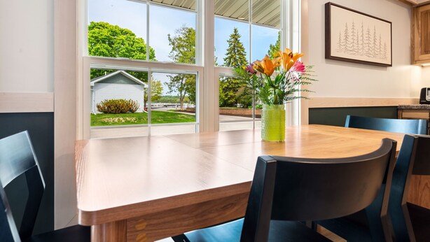 A dining area with a wooden table and chairs, a vase with flowers, and a view of greenery and a small building outside. - Spruce Suite | Downtown Fish Creek~Dog-friendly (Fish Creek)