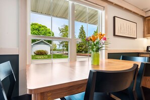 Uma sala de jantar com mesa e cadeiras de madeira, um vaso com flores e vista para o verde e um pequeno prédio lá fora.