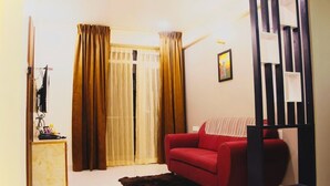 A living room with a red sofa, a small wooden cabinet, a black and white shelf unit, and a large window with curtains. - Paradise Point Ooty (Coonoor)