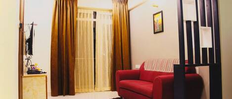 Una sala de estar con un sofá rojo, un mueble de madera pequeño, una estantería blanco y negro y un ventanal con cortinas.