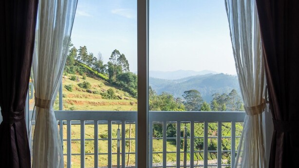 A view through a window with white curtains, revealing a balcony with a railing and a landscape of green hills and trees. - Paradise Point Ooty (Coonoor)