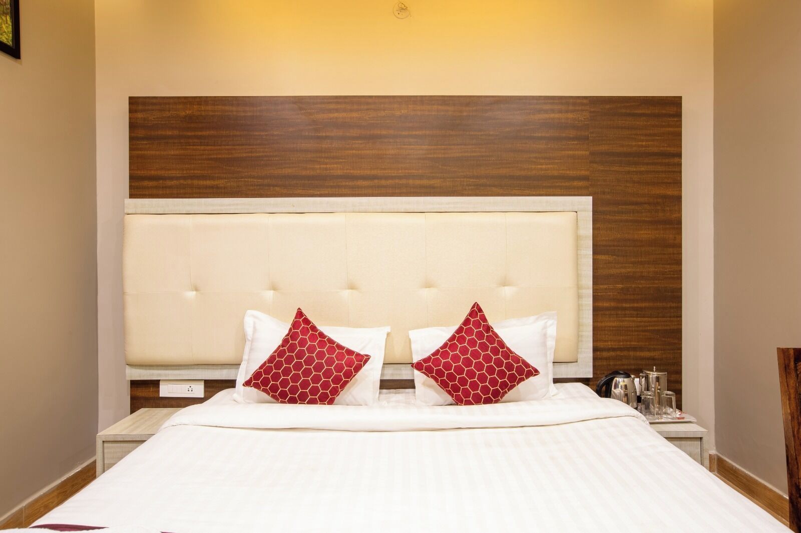 A hotel room with a large bed, two red patterned pillows, a wooden headboard, and a nightstand with a lamp and a glass of water.