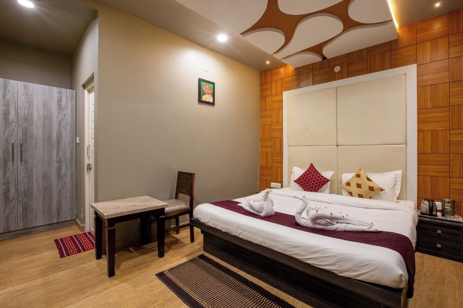 Een hotelkamer met een groot bed, een houten garderobe, een kleine tafel met stoel, en een rode vloerkleed.