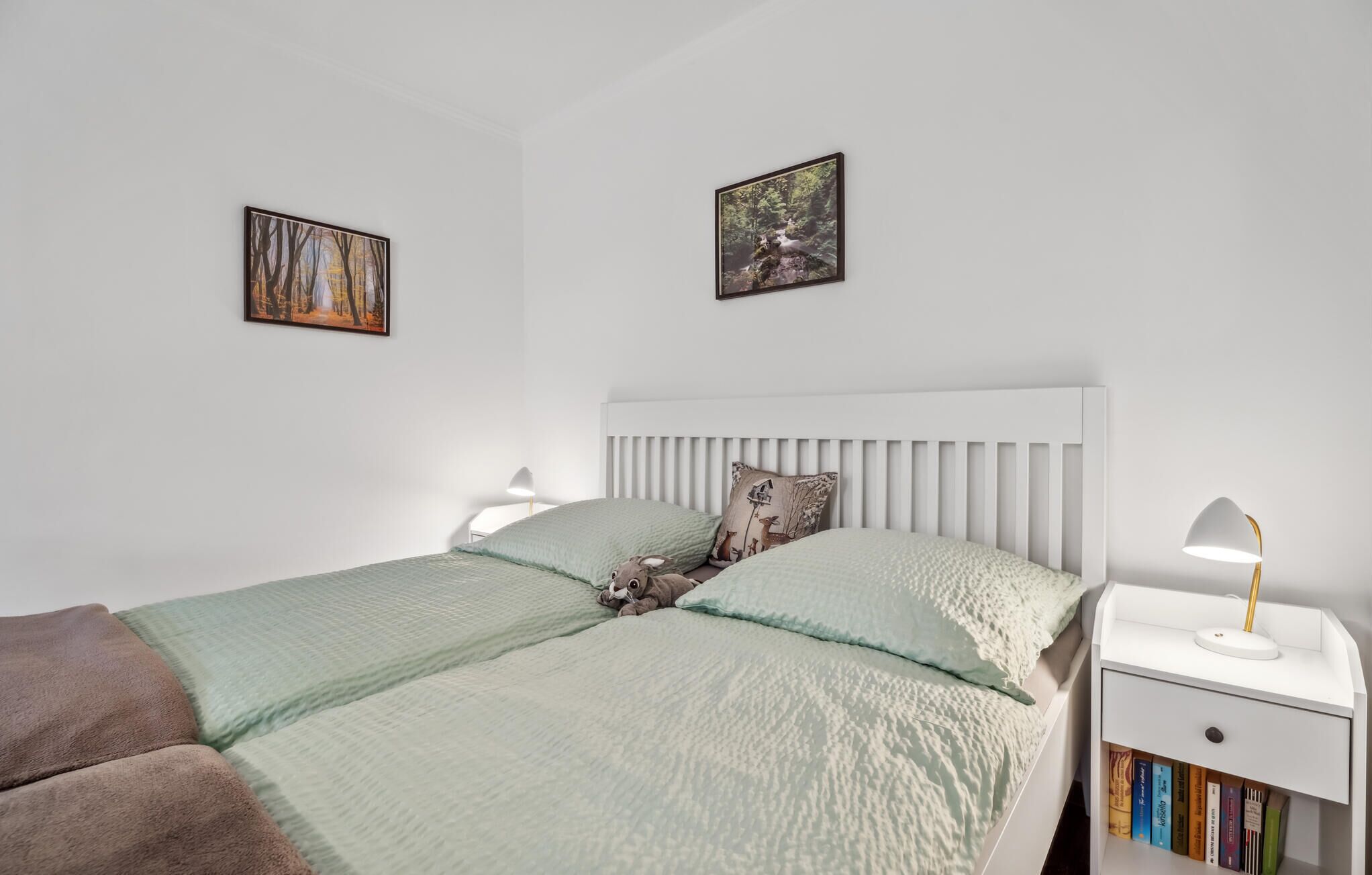 Een slaapkamer met een wit bed, twee kussens, een nachtkastje met een lamp en twee ingelijste foto's aan de muur.