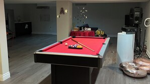 Une table de billard dans une pièce dotée d’un plancher en bois, d’une kitchenette et d’un coin salon avec un fauteuil.