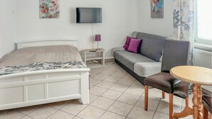 Une chambre à coucher avec un lit, un canapé, une télévision, une petite table et une fenêtre avec des rideaux.