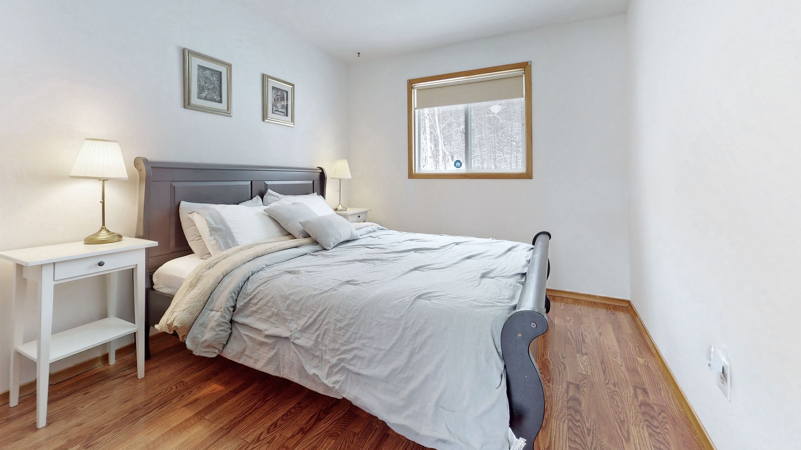 A bedroom with a bed, two bedside tables, a nightstand, a window with a view, and two framed pictures on the wall.