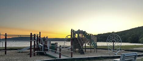 Een speeltuin met diverse speeltoestellen, een bankje en een uitzicht op een meer bij zonsondergang.
