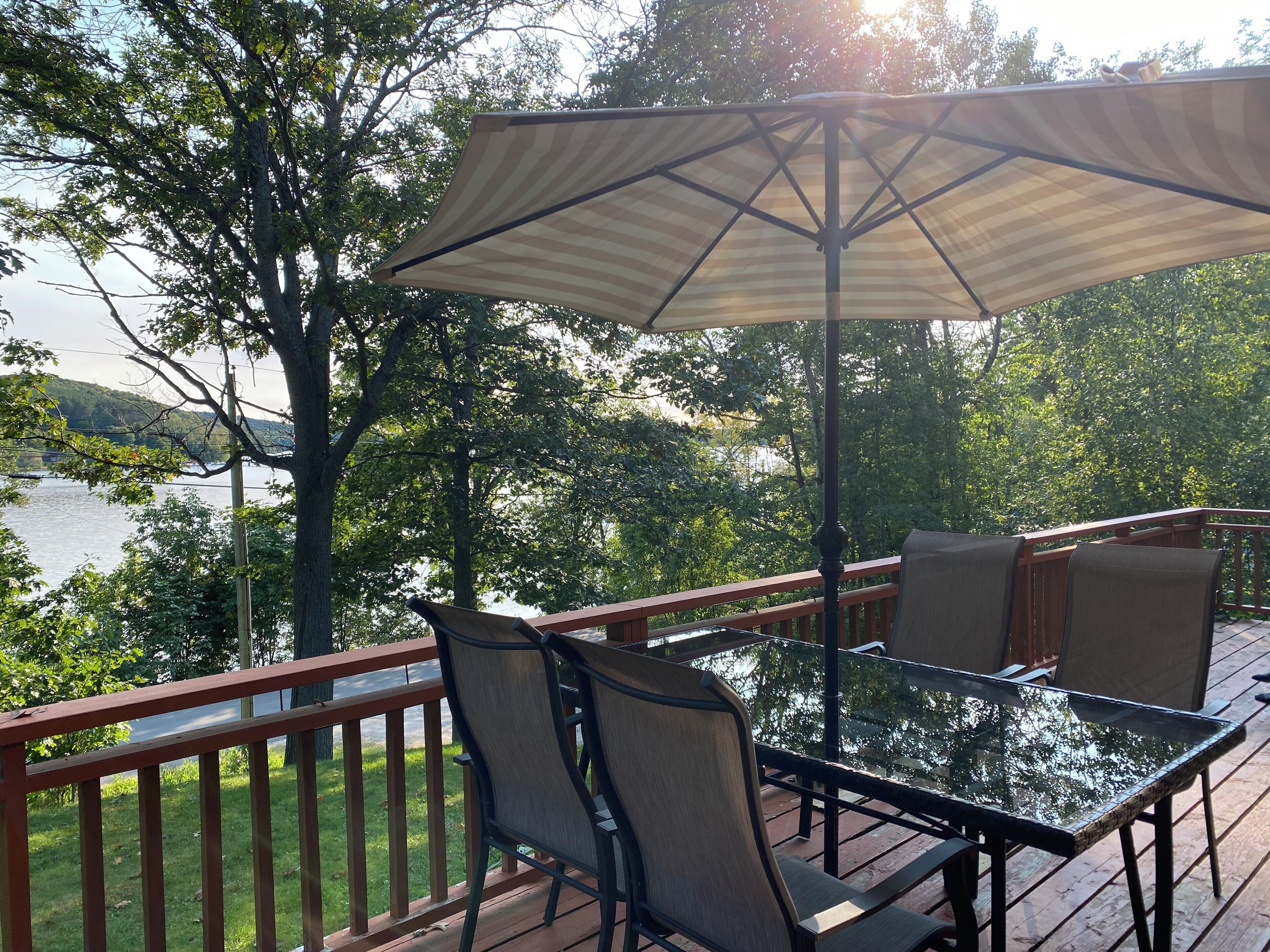 Une terrasse avec une table et des chaises en verre, un parasol à rayures et une vue sur un lac et des arbres.