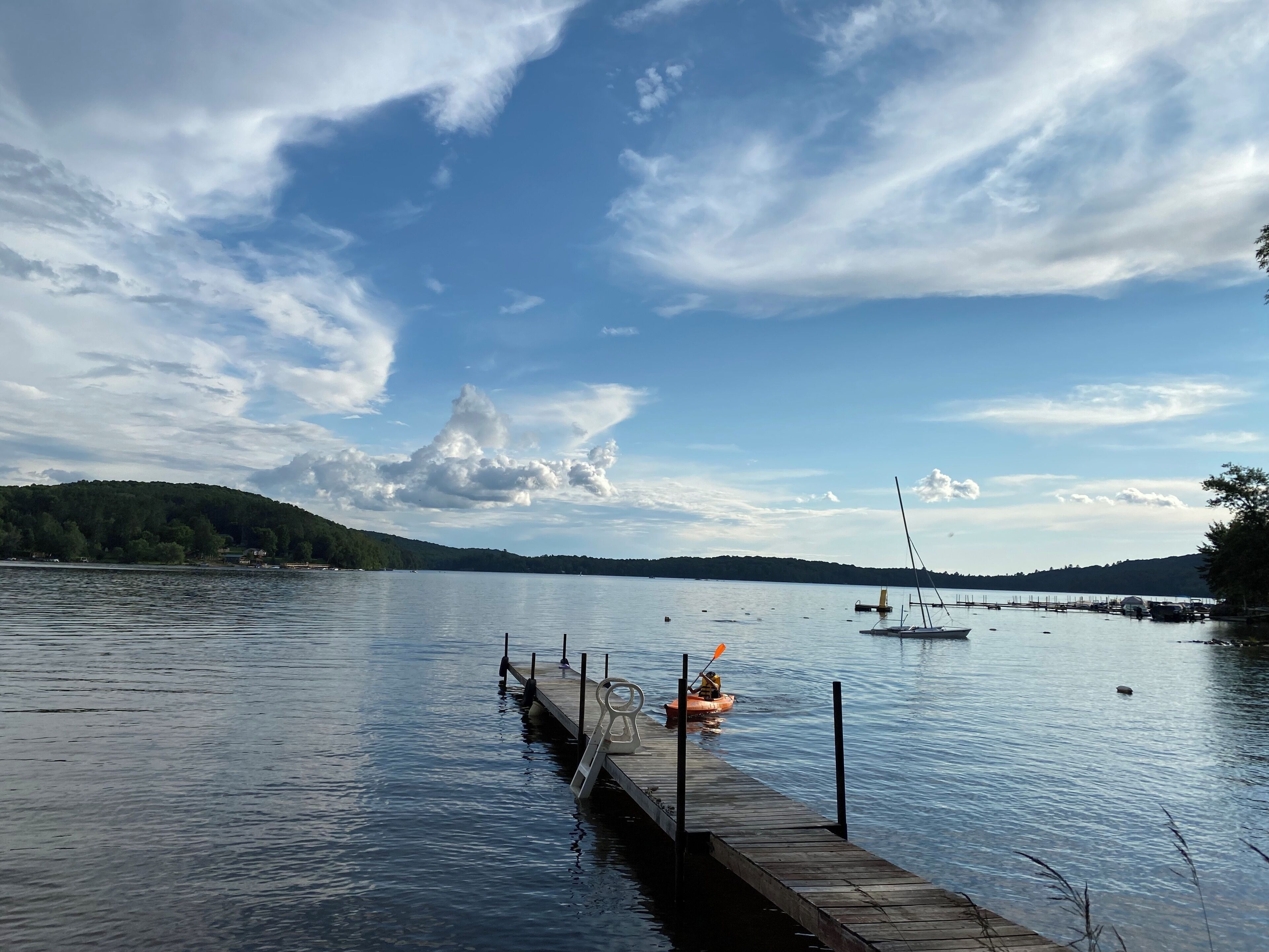 A calm lake with a wooden dock extending into the water, a kayak, and a sailboat in the distance.