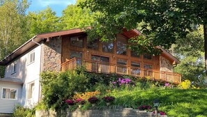 A two-story wooden house with a stone facade, a wooden balcony, and a lush garden with various flowers. - Charming lakefront cottage on Little Doe Lake in Muskoka area (Katrine)