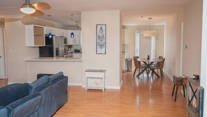 A modern living room with a blue sofa, a dining area with a wooden table and chairs, and a kitchen with white cabinets and stainless steel appliances.
