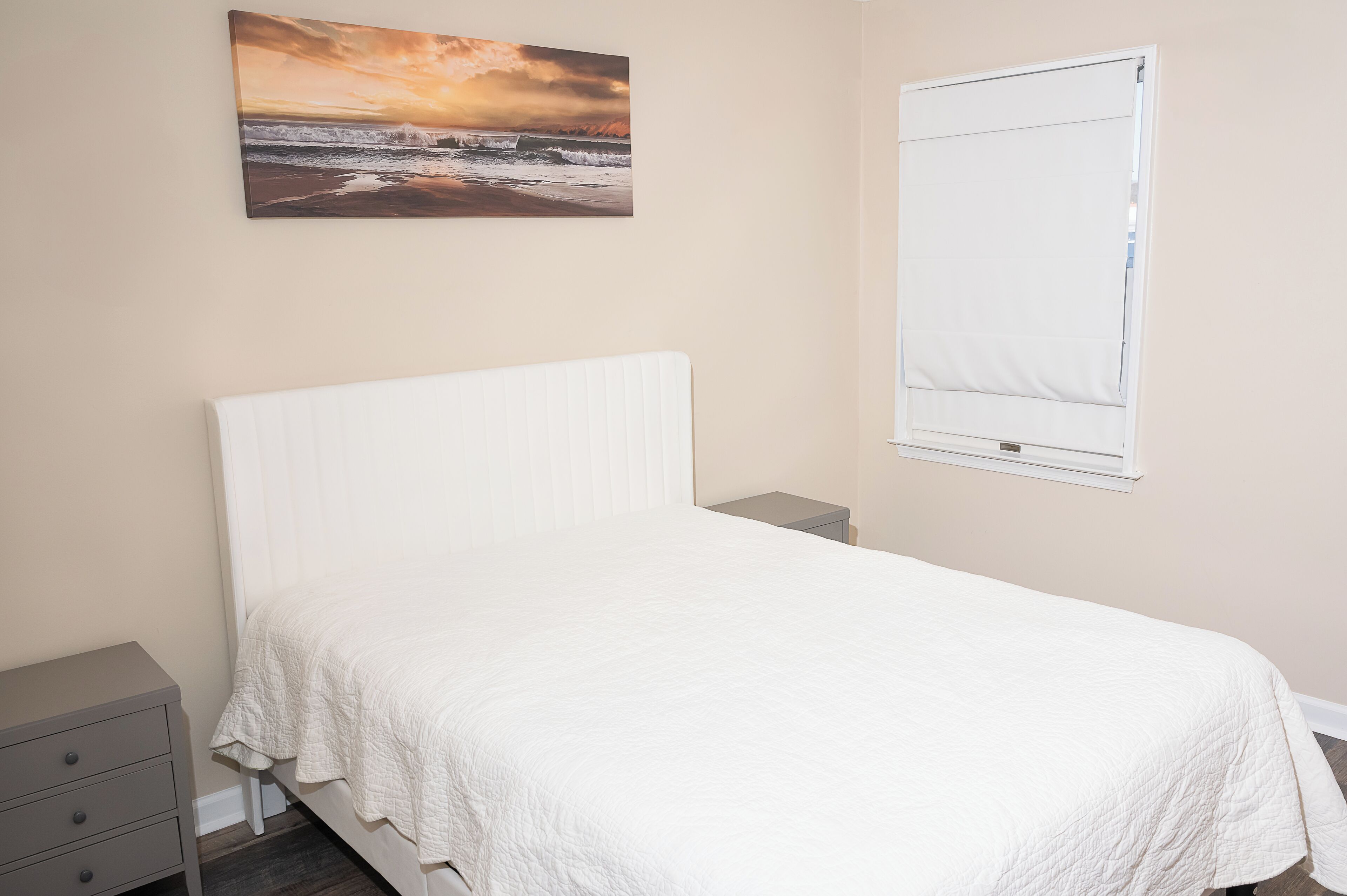 A bedroom with a bed, a nightstand, a drawer, a window with blinds, and a painting on the wall.