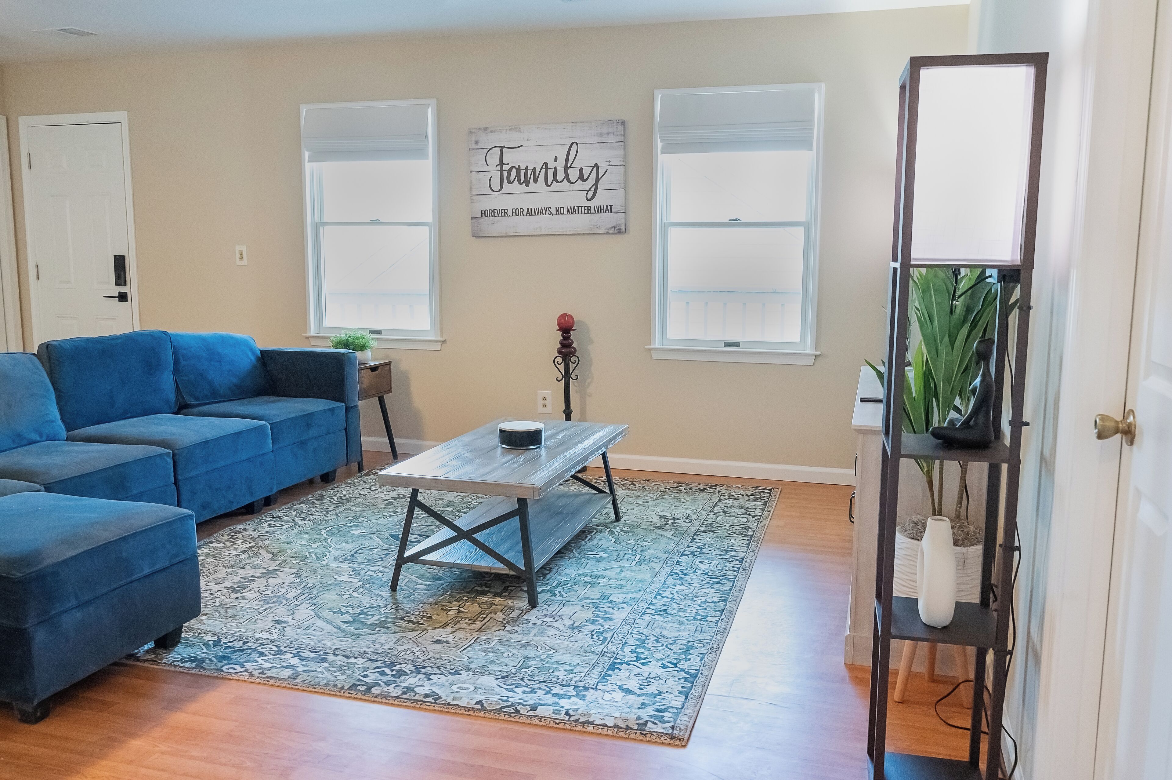 A living room with a blue sofa, a coffee table, a rug, and a wall sign that reads 'Family Forever, For Always, No Matter What.'