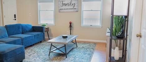 A living room with a blue sofa, a coffee table, a rug, and a wall sign that reads 'Family Forever, For Always, No Matter What.'